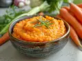 Un bol de purée de carotte onctueuse, garni d'une branche de romarin, avec des carottes fraîches en arrière-plan.