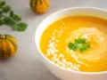 Un bol de soupe butternut crémeuse, garnie de coriandre fraîche et de petits morceaux de fromage. Des mini-citrouilles décorent la table.