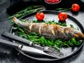 Un bar entier, parfaitement doré, est prêt pour la dégustation. Ce plat de cuisson bar au four est servi sur un lit de roquette fraîche, accompagné de tomates cerises et de romarin.