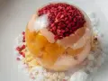 Sphère transparente garnie de fruits rouges et de pêche melba, sur un lit de miettes et de glace pilée.