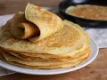 Pile de crêpes dorées, une roulée sur le dessus. Une poêle avec une crêpe en cuisson est visible en arrière-plan. Parfait pour 1 litre de pâte à crêpe.