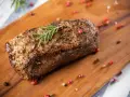 Un délicieux roti de boeuf, assaisonné et garni de romarin, repose sur une planche en bois, entouré de grains de poivre rose et noir.