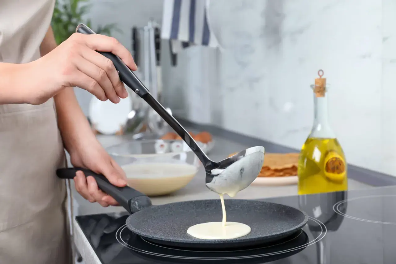 Une louche verse de la pâte dans une poêle pour une recette pâte à crêpe facile. Huile d'olive et pile de crêpes en arrière-plan.