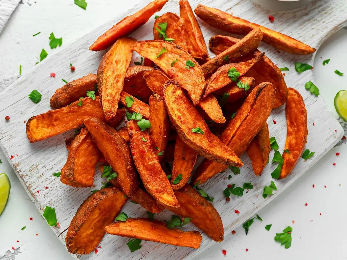 Des frites de patate douce dorées et croustillantes, parsemées de persil frais et de poivre rose, sur une planche en bois.