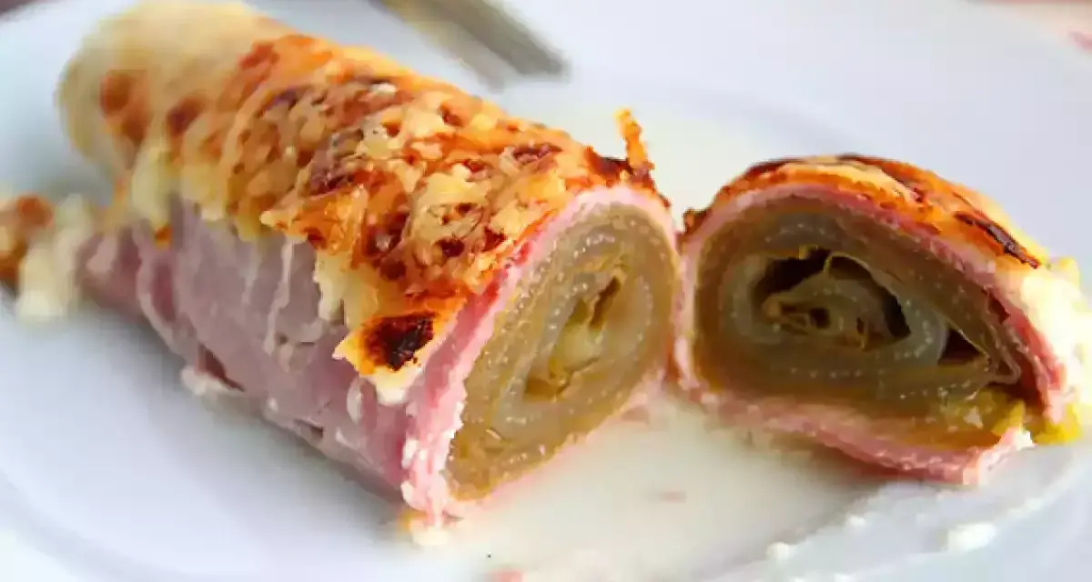 Endive au jambon gratinée, enroulée et coupée en deux, servie sur une assiette blanche avec une sauce crémeuse.