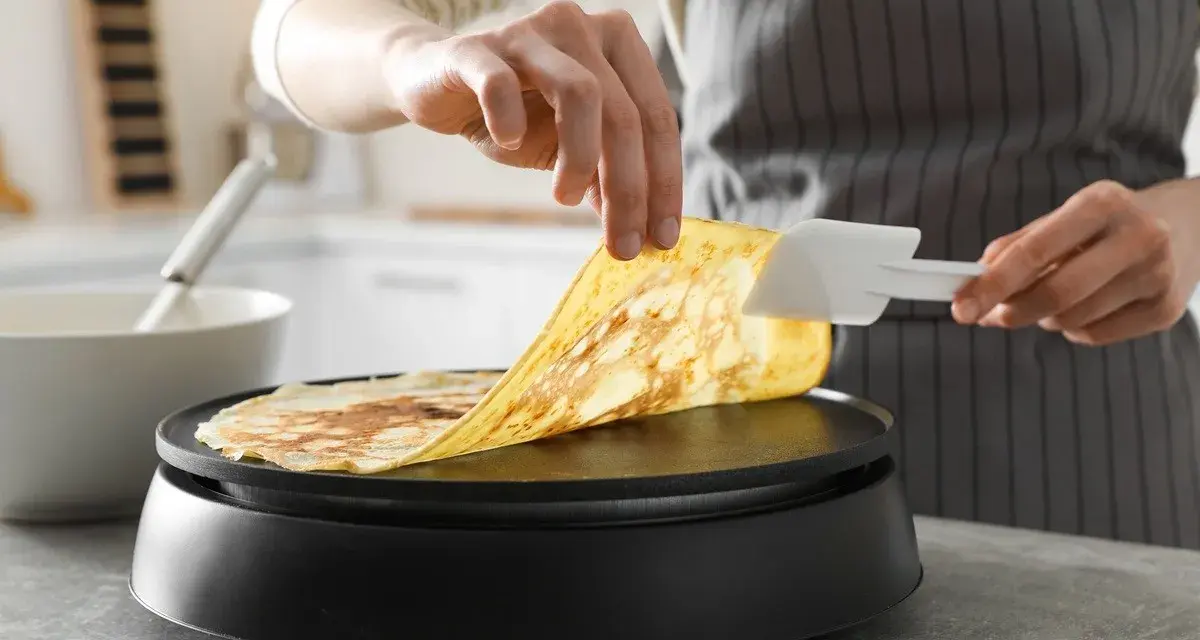 Une personne utilise une spatule pour soulever une crêpe dorée de la plaque chauffante. Astuce pour comment nettoyer une crêpière électrique.