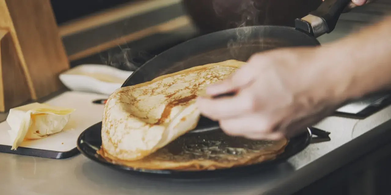 Une main retourne une crêpe dorée dans une poêle fumante. Sur le côté, du beurre frais attend. Ces meilleures crêpes promettent un délice.