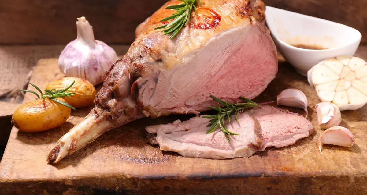 Gigot d'agneau rôti, parfait pour une recette d'agneau pascal. Accompagné de pommes de terre, ail et romarin sur une planche en bois.
