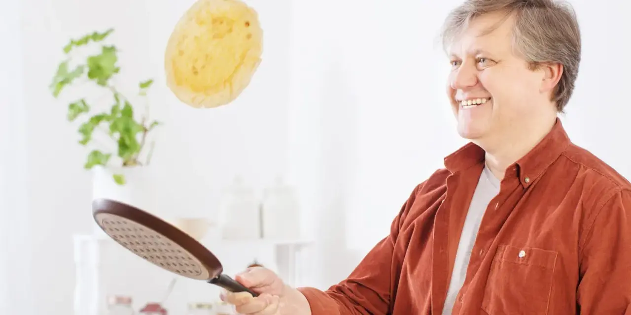 Un homme souriant s'amuse à retourner une crêpe en plein vol avec une poêle.