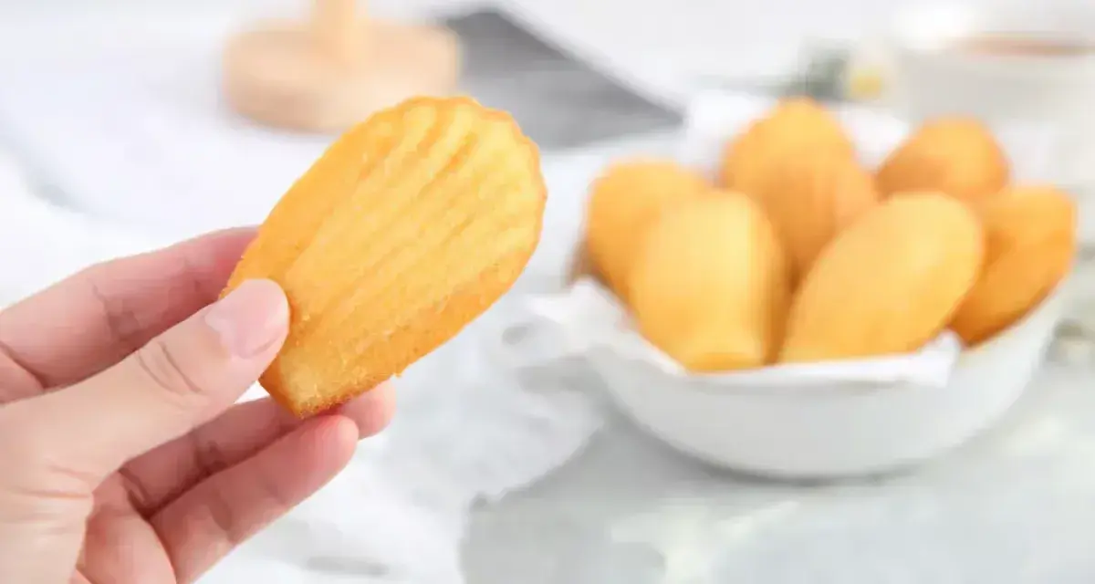 Une main tient une madeleine dorée, parfaite pour une recette madeleine moelleuse. Un bol rempli de ces délicieuses pâtisseries est visible en arrière-plan.