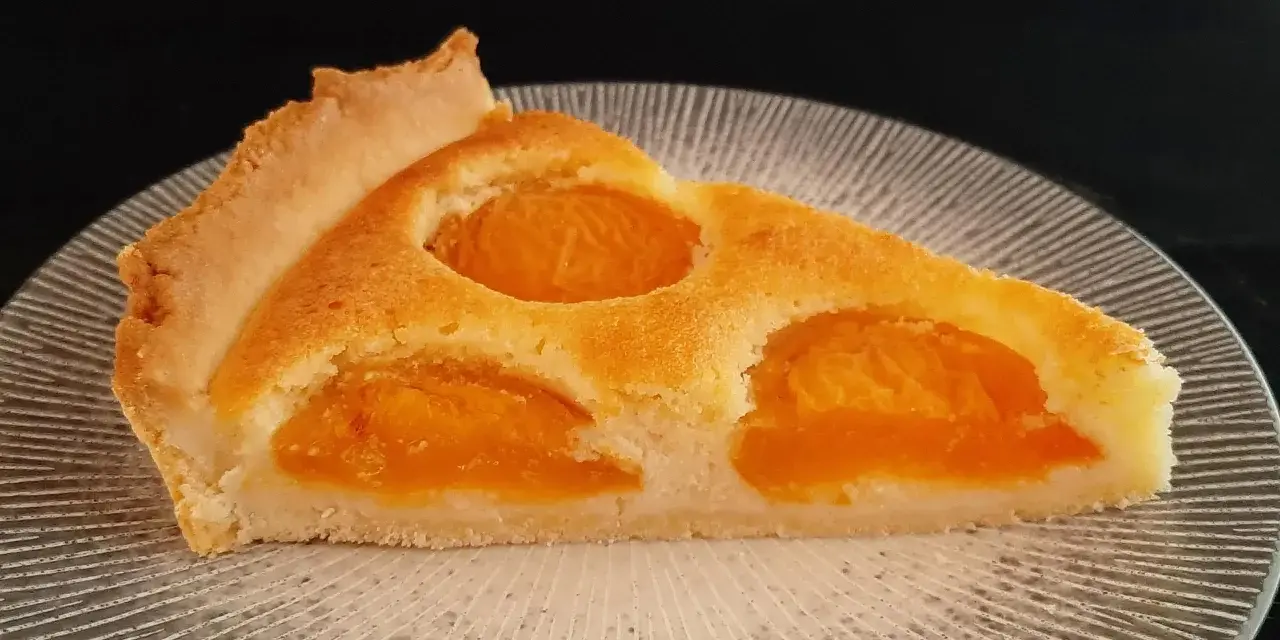 Une part de tarte aux abricots dorée, avec des fruits juteux incrustés dans une pâte moelleuse, sur une assiette texturée.