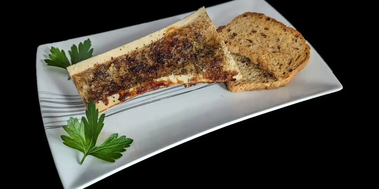 Un os à moelle rôti, assaisonné et garni de persil, servi avec des toasts grillés sur une assiette blanche.