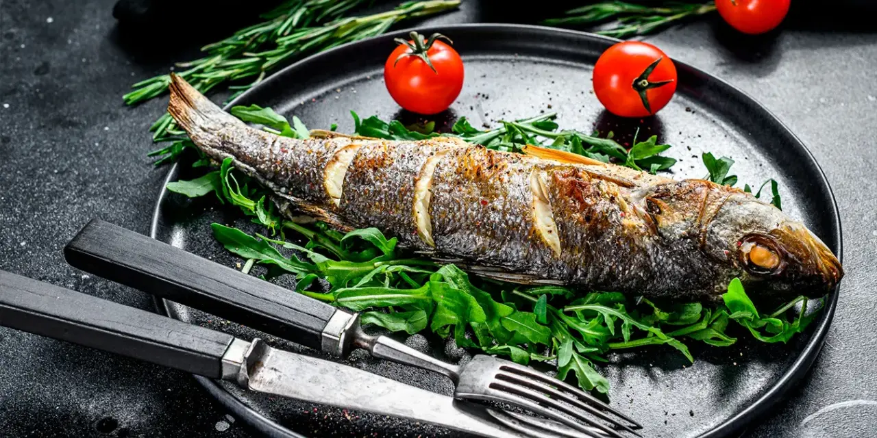 Un bar entier, parfaitement doré, est prêt pour la dégustation. Ce plat de cuisson bar au four est servi sur un lit de roquette fraîche, accompagné de tomates cerises et de romarin.