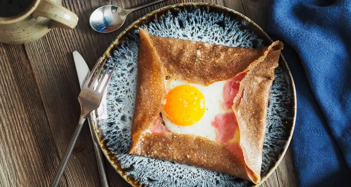 Une galette de sarrasin garnie d'un œuf au plat et de jambon, un des nombreux types de crêpes salées. Servi sur une assiette avec des couverts.