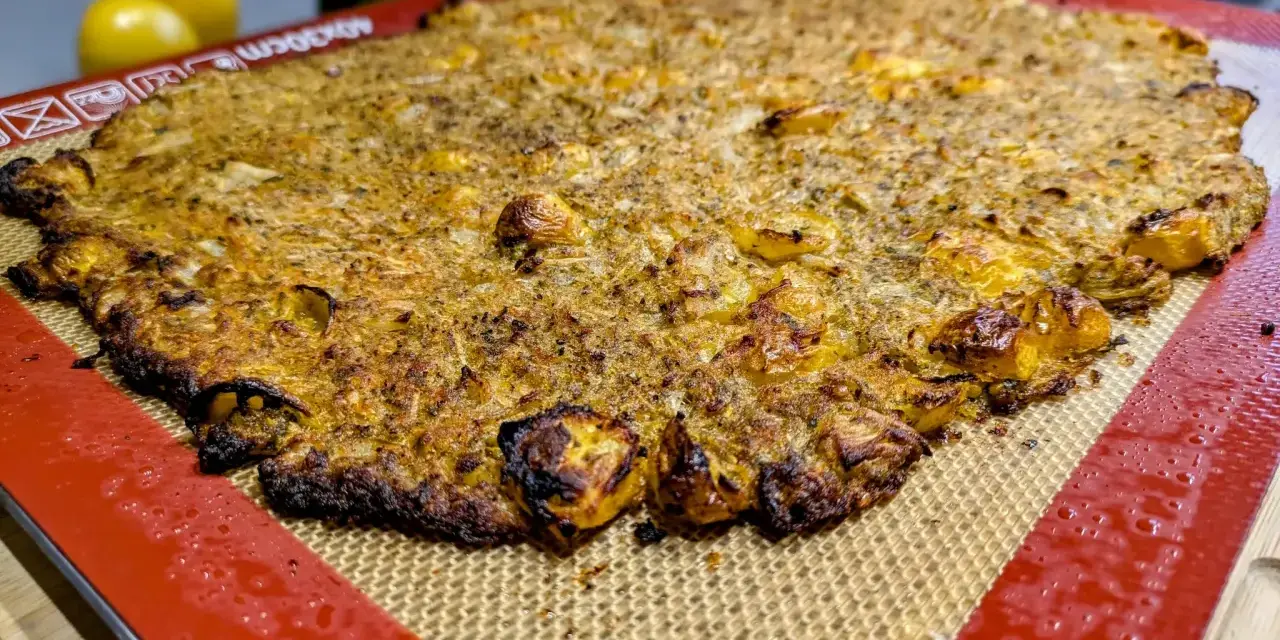 Une galette complète au four, dorée et croustillante, sur un tapis de cuisson en silicone. Des tomates jaunes et une courgette sont floues en arrière-plan.