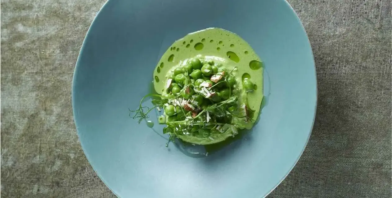 Délicieuse cuisson petit pois frais, servis dans une assiette bleue avec une émulsion verte et des éclats d'amandes.