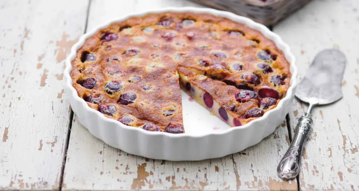 Un délicieux clafoutis aux cerises, encore chaud dans son plat blanc, avec une part coupée.