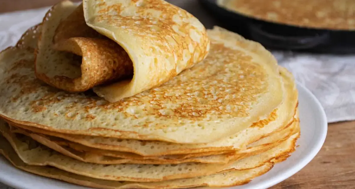 Pile de crêpes dorées, une roulée sur le dessus. Une poêle avec une crêpe en cuisson est visible en arrière-plan. Parfait pour 1 litre de pâte à crêpe.