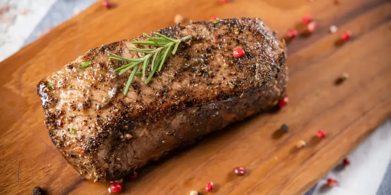 Un délicieux roti de boeuf, assaisonné et garni de romarin, repose sur une planche en bois, entouré de grains de poivre rose et noir.