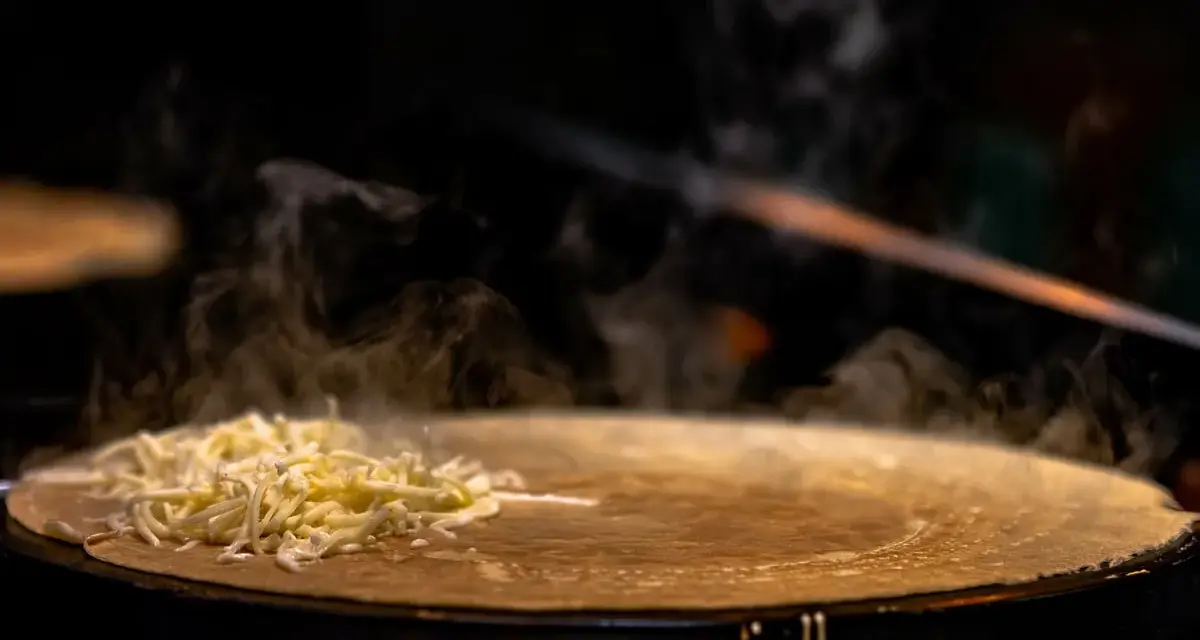 Fromage râpé fondant sur une crêpe chaude, vapeur s'élevant. Une tâche de nettoyage crêpière Tefal brûlée est visible sur le bord.