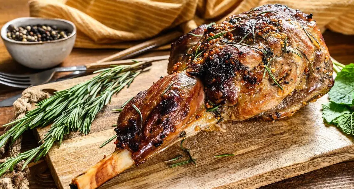 Selle d'agneau rôtie, garnie de romarin et de moutarde, sur une planche en bois avec des herbes fraîches.
