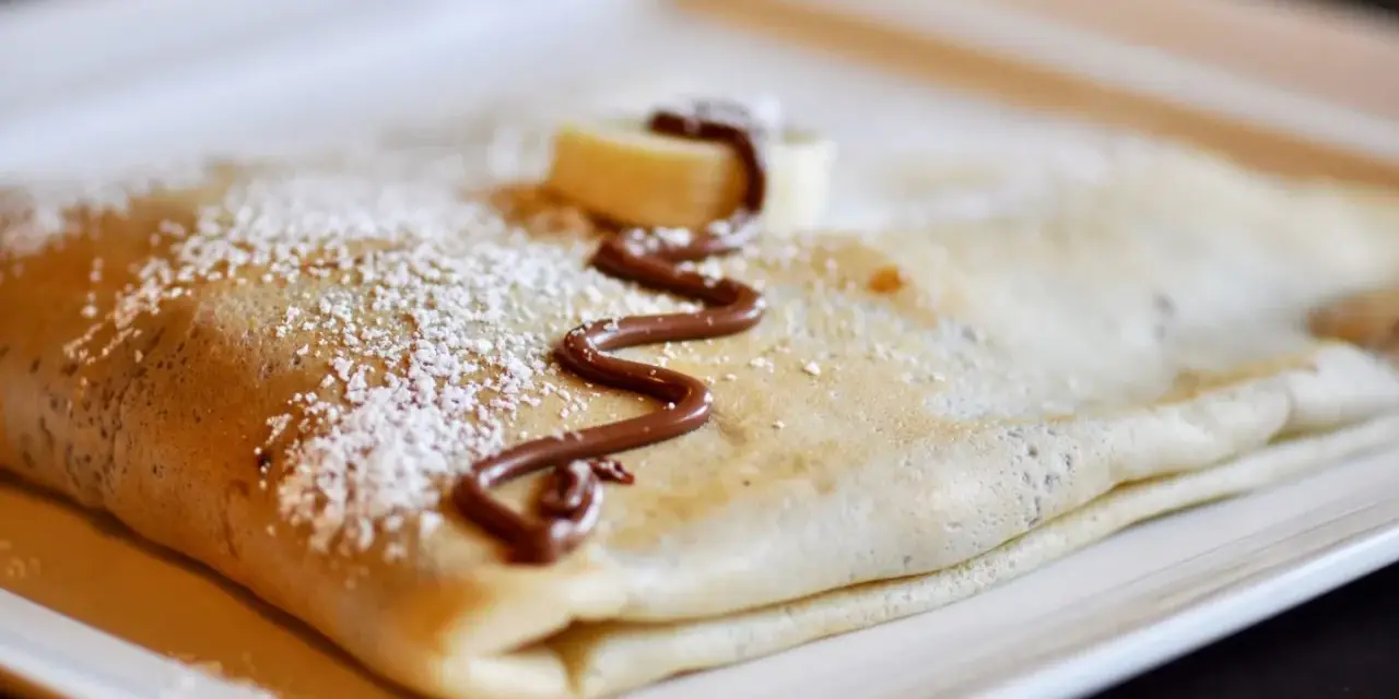 Crêpe pliée, saupoudrée de sucre glace, décorée de chocolat fondu et d'une rondelle de banane. La meilleure recette de pâte à crêpes pour un délice !