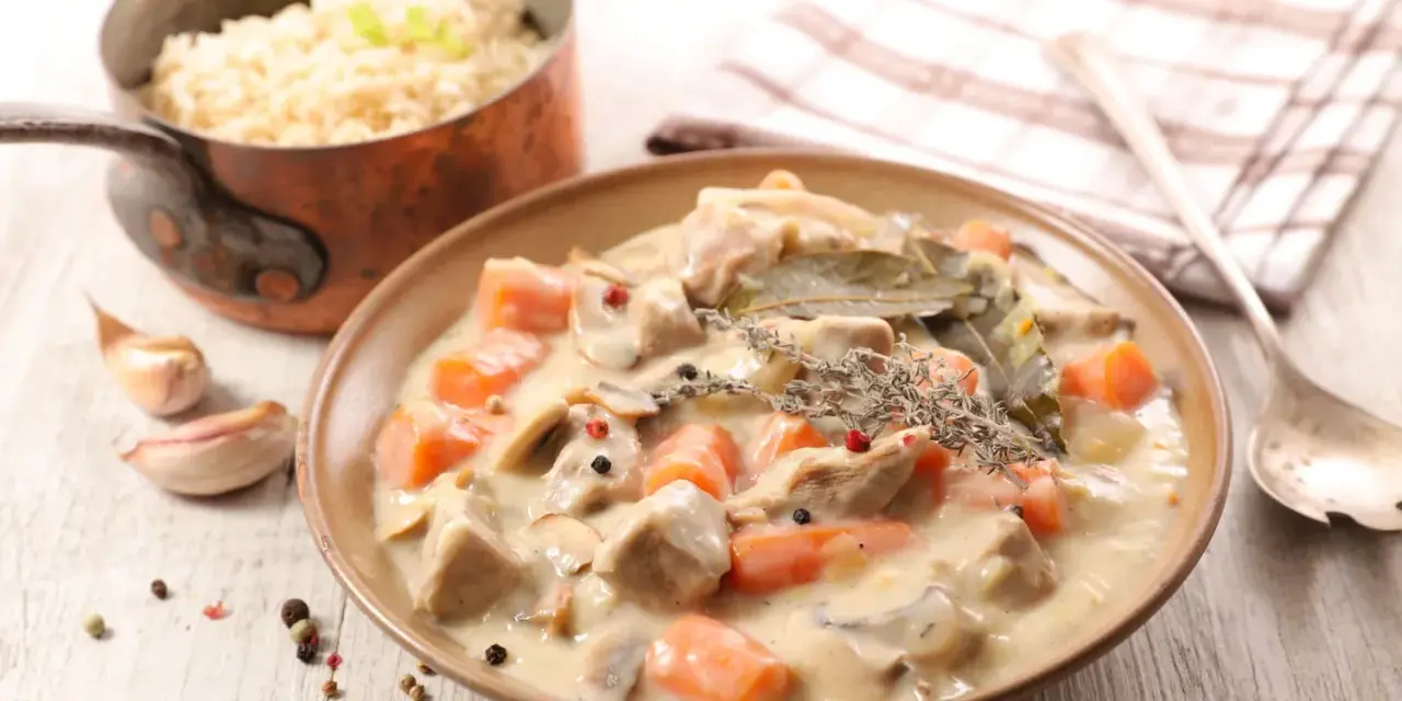 Un plat de blanquette de veau crémeuse avec des carottes, des champignons et des herbes, servi avec du riz dans une casserole en cuivre.