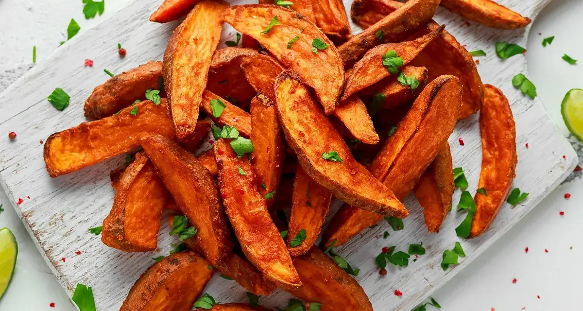 Des frites de patate douce dorées et croustillantes, parsemées de persil frais et de poivre rose, sur une planche en bois.