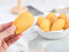 Une main tient une madeleine dorée, parfaite pour une recette madeleine moelleuse. Un bol rempli de ces délicieuses pâtisseries est visible en arrière-plan.