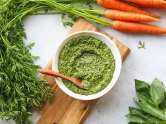 Un bol de pesto de fanes de carottes, avec des carottes fraîches et du basilic à côté.