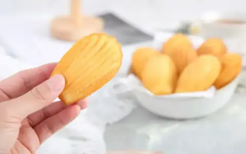 Une main tient une madeleine dorée, parfaite pour une recette madeleine moelleuse. Un bol rempli de ces délicieuses pâtisseries est visible en arrière-plan.