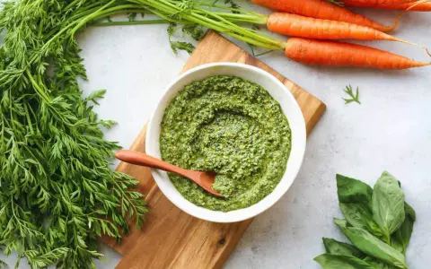 Un bol de pesto de fanes de carottes, avec des carottes fraîches et du basilic à côté.