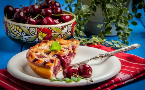Une part de tarte aux cerises, garnie de menthe fraîche, sur une assiette blanche avec une fourchette. Un bol de cerises est visible en arrière-plan.