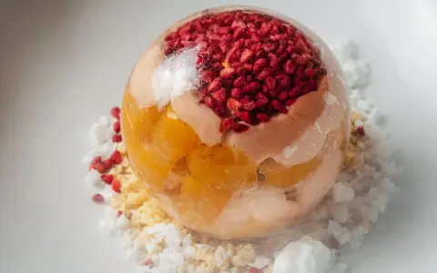 Sphère transparente garnie de fruits rouges et de pêche melba, sur un lit de miettes et de glace pilée.