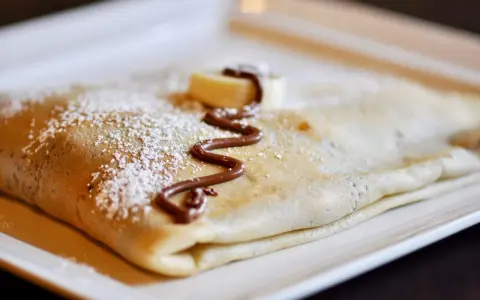 Crêpe pliée, saupoudrée de sucre glace, décorée de chocolat fondu et d'une rondelle de banane. La meilleure recette de pâte à crêpes pour un délice !
