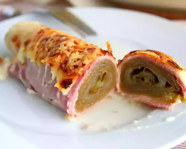 Endive au jambon gratinée, enroulée et coupée en deux, servie sur une assiette blanche avec une sauce crémeuse.