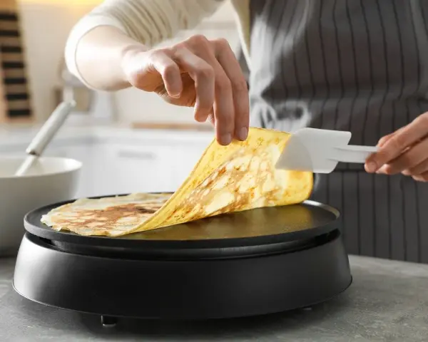 Une personne utilise une spatule pour soulever une crêpe dorée de la plaque chauffante. Astuce pour comment nettoyer une crêpière électrique.