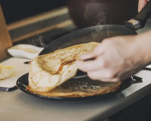 Une main retourne une crêpe dorée dans une poêle fumante. Sur le côté, du beurre frais attend. Ces meilleures crêpes promettent un délice.