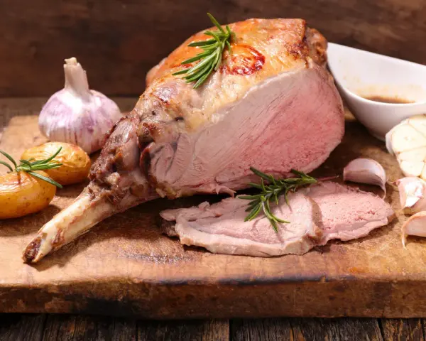 Gigot d'agneau rôti, parfait pour une recette d'agneau pascal. Accompagné de pommes de terre, ail et romarin sur une planche en bois.