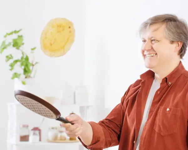 Un homme souriant s'amuse à retourner une crêpe en plein vol avec une poêle.