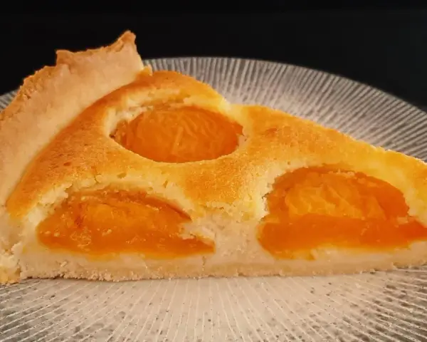 Une part de tarte aux abricots dorée, avec des fruits juteux incrustés dans une pâte moelleuse, sur une assiette texturée.