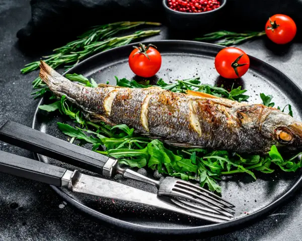 Un bar entier, parfaitement doré, est prêt pour la dégustation. Ce plat de cuisson bar au four est servi sur un lit de roquette fraîche, accompagné de tomates cerises et de romarin.