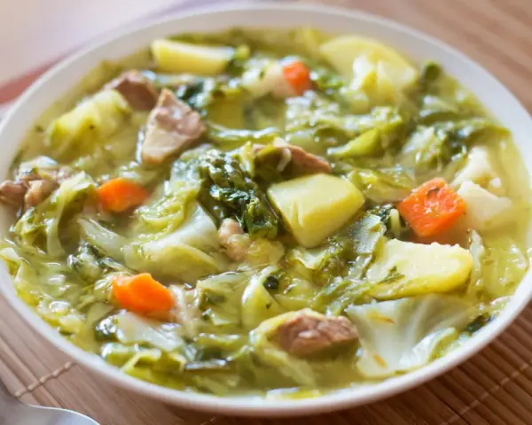 Un bol de soupe aux choux réconfortante, avec des morceaux de chou, pommes de terre, carottes et viande. Du pain frais à côté.