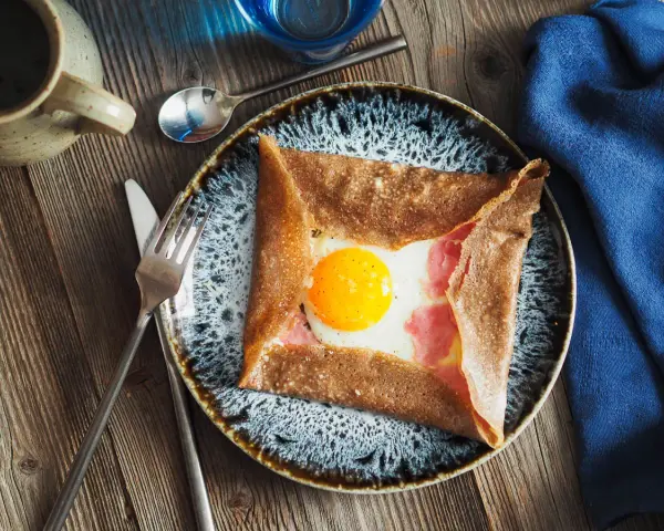 Une galette de sarrasin garnie d'un œuf au plat et de jambon, un des nombreux types de crêpes salées. Servi sur une assiette avec des couverts.