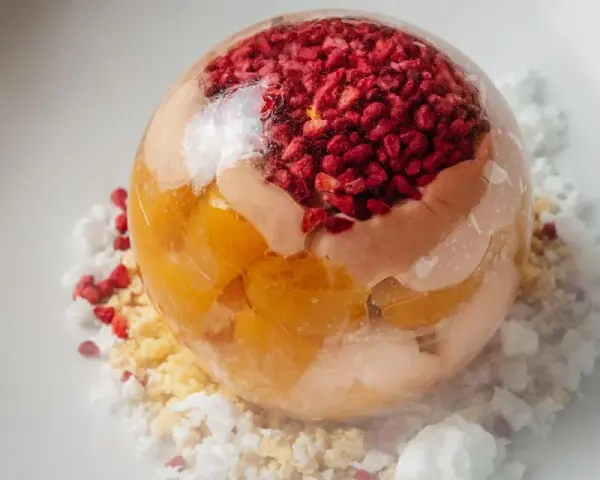 Sphère transparente garnie de fruits rouges et de pêche melba, sur un lit de miettes et de glace pilée.