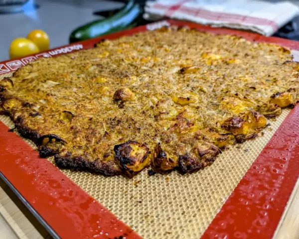Une galette complète au four, dorée et croustillante, sur un tapis de cuisson en silicone. Des tomates jaunes et une courgette sont floues en arrière-plan.