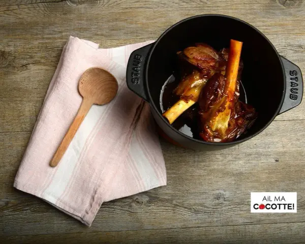Souris d'agneau à la cocotte en fonte, tendres et savoureuses, prêtes à être dégustées. Une cuillère en bois et une serviette rose complètent la scène.