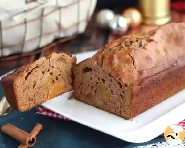 Un délicieux pain d'épices aux fruits confits, coupé en deux, sur une assiette blanche. Des bâtons de cannelle et des décorations de Noël en arrière-plan.