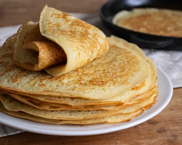 Pile de crêpes dorées, une roulée sur le dessus. Une poêle avec une crêpe en cuisson est visible en arrière-plan. Parfait pour 1 litre de pâte à crêpe.