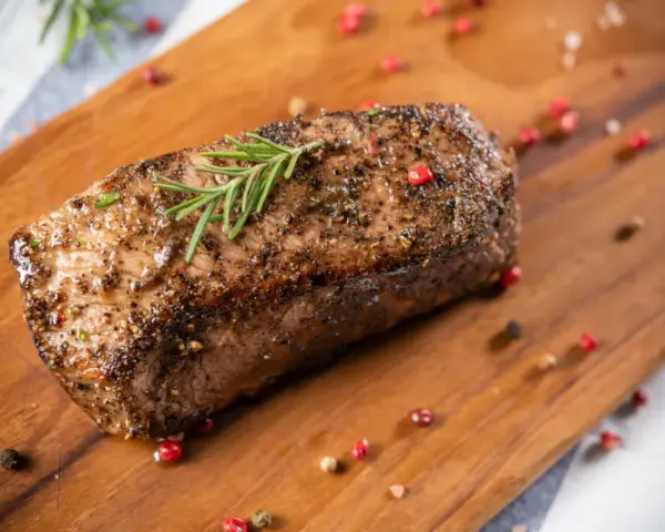 Un délicieux roti de boeuf, assaisonné et garni de romarin, repose sur une planche en bois, entouré de grains de poivre rose et noir.